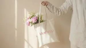 Brazo sosteniendo una bolsa de tela con el logotipo de Morocco Beauty y un ramo de flores frescas, en una composición horizontal con luz solar cálida