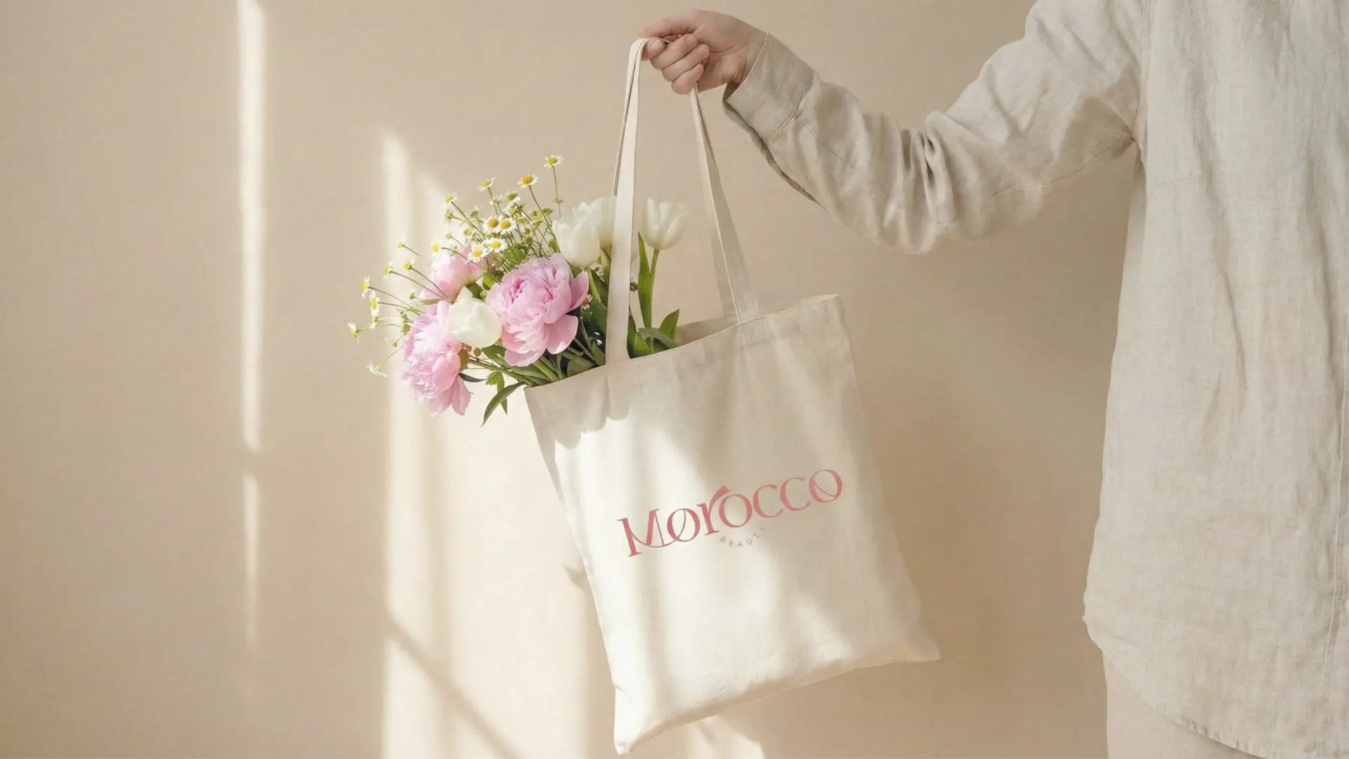 Brazo sosteniendo una bolsa de tela con el logotipo de Morocco Beauty y un ramo de flores frescas, en una composición horizontal con luz solar cálida