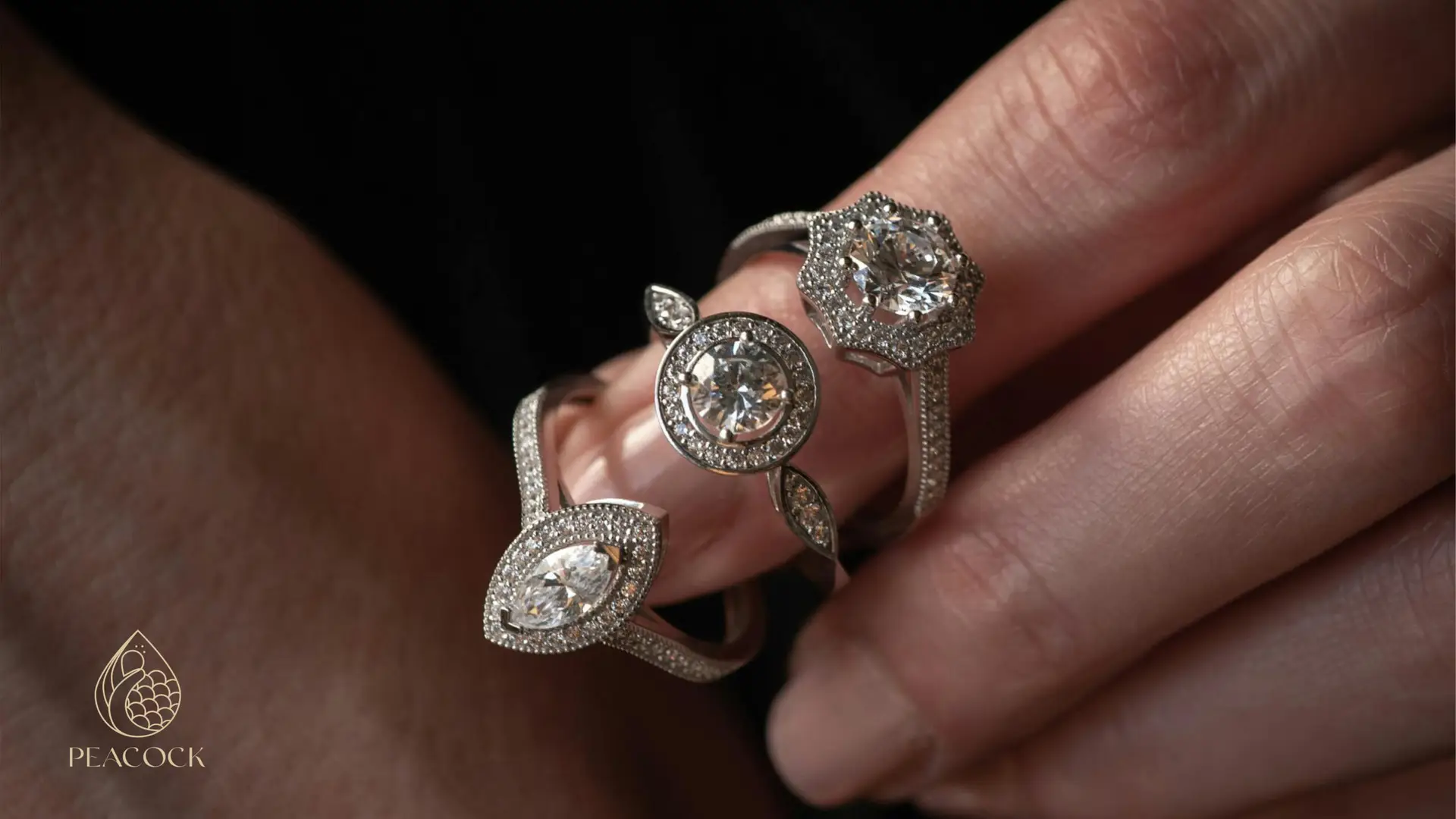 Mano con tres anillos de diamantes de lujo en primer plano y el logotipo de Peacock Fine Jewelry en la esquina inferior izquierda.
