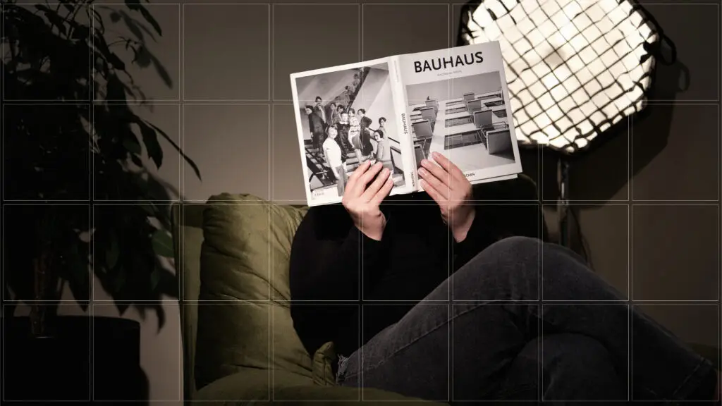 Persona sentada en un sofá verde oscuro sosteniendo un libro abierto de la Bauhaus frente a su rostro, en una habitación con iluminación de estudio y fondo oscuro.