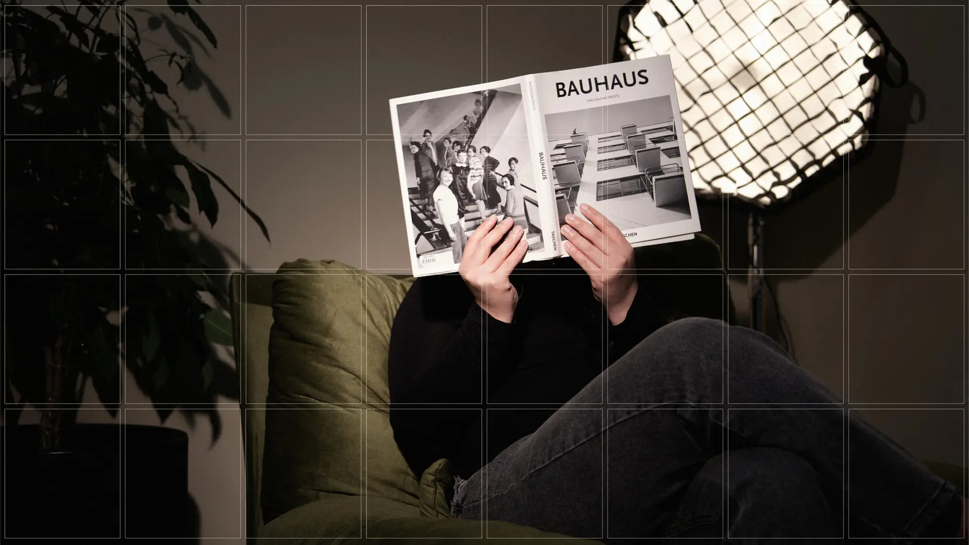 Persona sentada en un sofá verde oscuro sosteniendo un libro abierto de la Bauhaus frente a su rostro, en una habitación con iluminación de estudio y fondo oscuro.